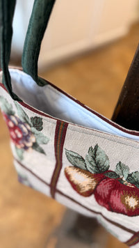 A close-up of a woven tote bag with dark green handles, featuring a cream-colored body with embroidered red apples, green leaves, and a brown plaid pattern, hanging near a dark wooden post.