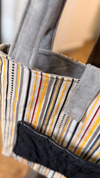 Close-up of a striped fabric tote bag with gray handles and a black front pocket, hanging indoors.