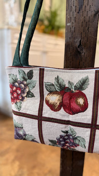 A close-up of a woven tote bag with a white background, featuring a grid pattern with red and green fruit motifs and dark green straps, hanging from a dark wooden post.