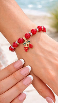 Red beaded bracelet with cherry charm on a wrist against a beach background