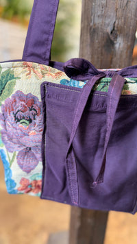 A purple fabric tote bag with a floral tapestry pattern side panel, hanging from a rustic wooden post. The bag features a solid purple front pocket with drawstring ties and a matching purple shoulder strap.