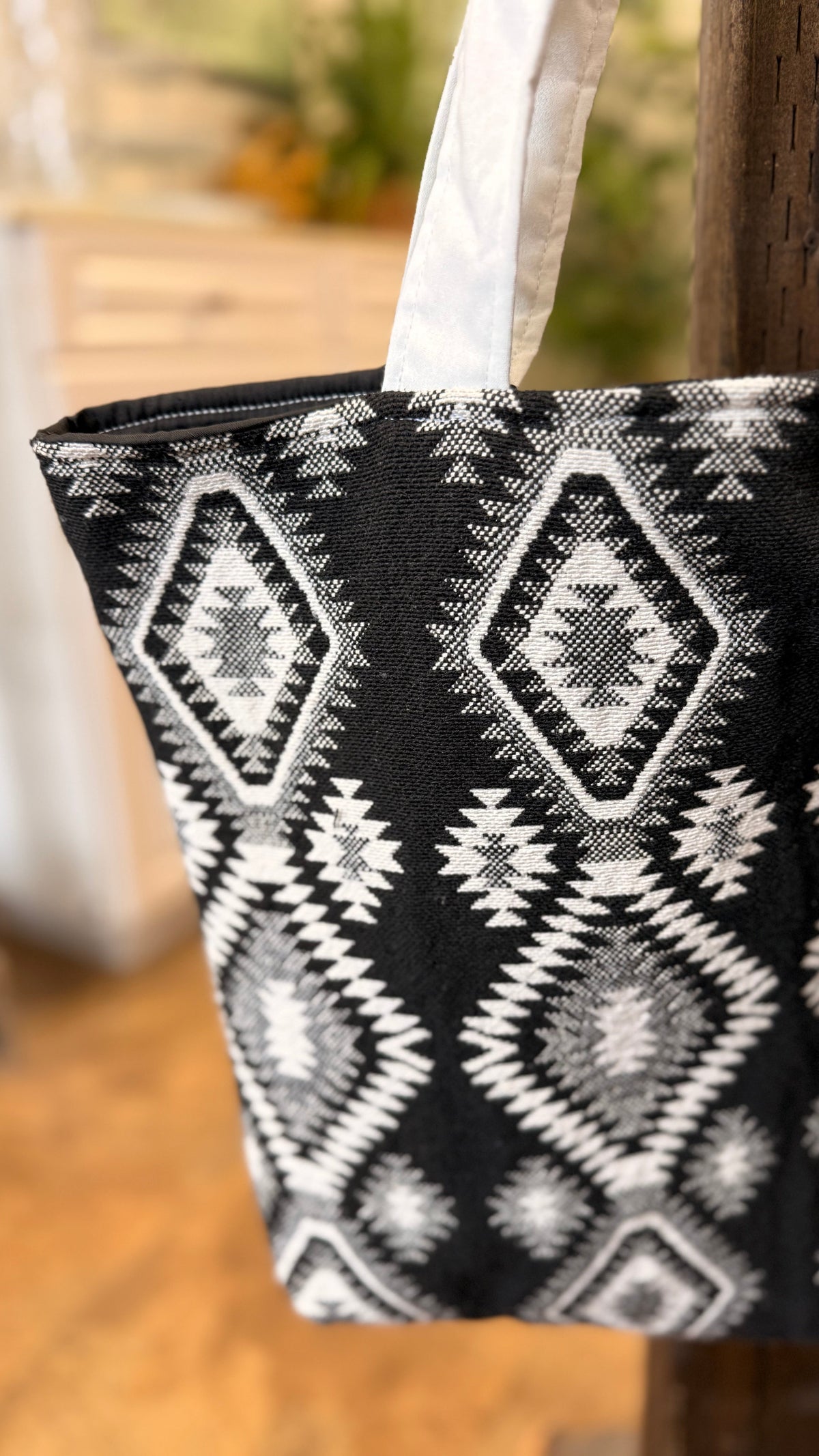 A close-up of a black tote bag with a white geometric diamond pattern, hanging from a white strap next to a wooden post in a softly blurred indoor setting.