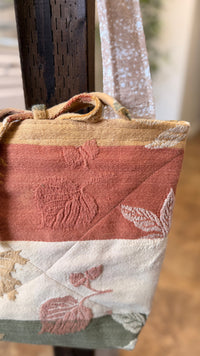 A close-up of a handmade quilted fabric tote bag hanging from a dark wooden chair. The bag features a patchwork design with horizontal stripes of dusty rose, cream, beige, and sage green, decorated with embroidered floral and leaf motifs.