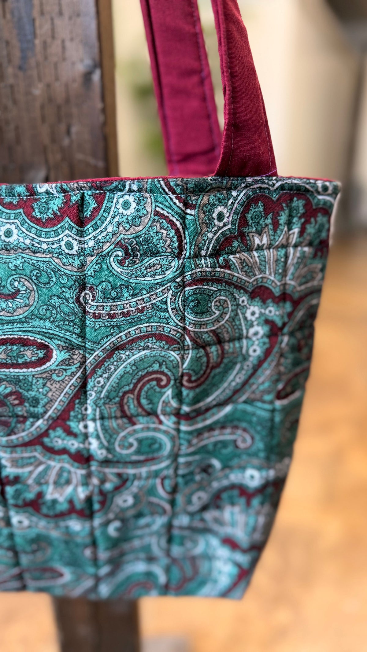 A close-up of a quilted fabric tote bag with a teal, burgundy, and white paisley pattern, hanging from a rustic wooden post.