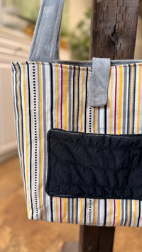 A striped fabric tote bag with gray shoulder straps and a black quilted front pocket, hanging on a rustic wooden stand in a softly blurred indoor setting.