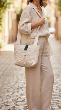 A cream-colored quilted tote bag with a dark green leather accent strap, hanging from a rustic wooden peg in a home interior setting.