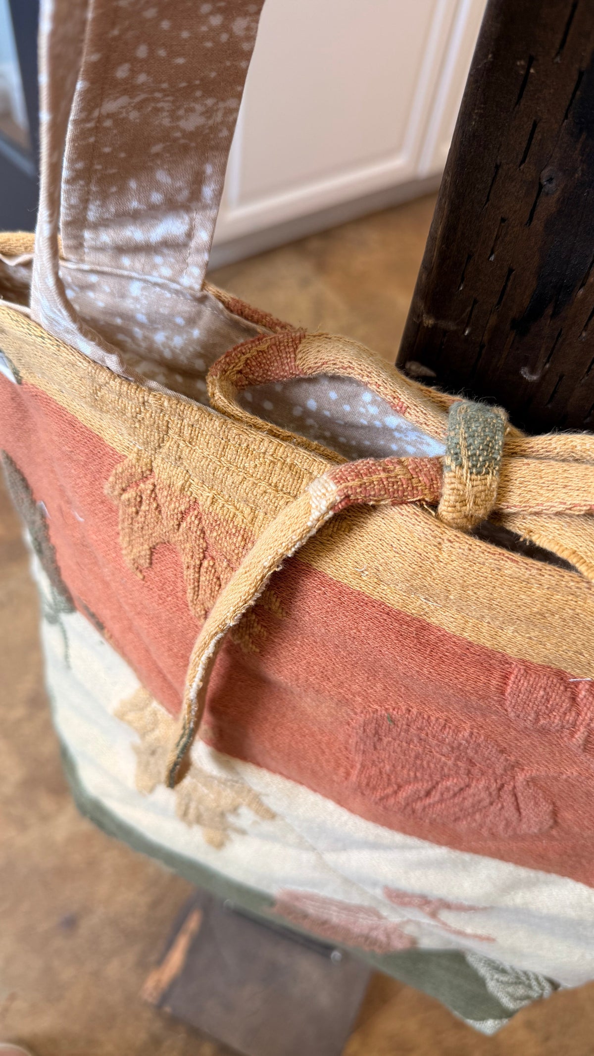 A close-up of a fabric tote bag with a rustic, distressed texture. The bag features a multi-colored pattern with horizontal bands of terracotta red, mustard yellow, cream, and muted green, accented by a matching woven strap tied around the opening.